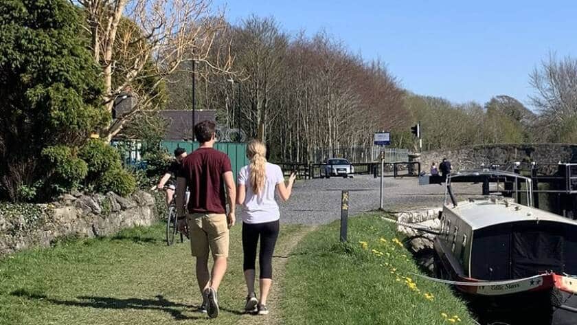 Two people walk along a grassy path beside a canal with a moored boat, while others walk and cycle ahead. Leafy trees and a stone wall line the path on a sunny day.