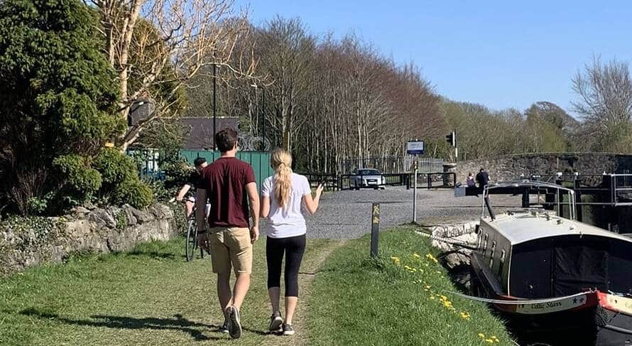 Two people walk along a grassy path beside a canal with a moored boat, while others walk and cycle ahead. Leafy trees and a stone wall line the path on a sunny day.