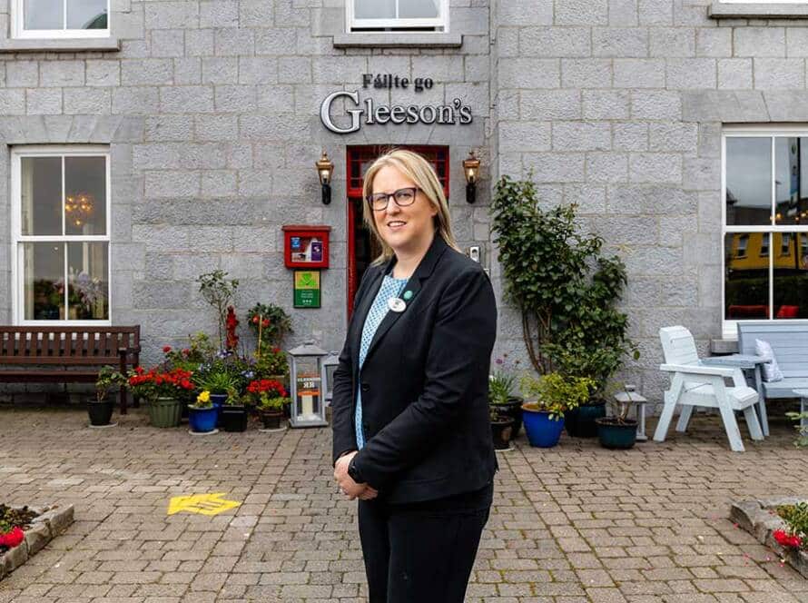 A woman in a black suit stands smiling outside Gleeson's, a grey stone building with potted plants, benches, and a sign in Irish above the entrance.