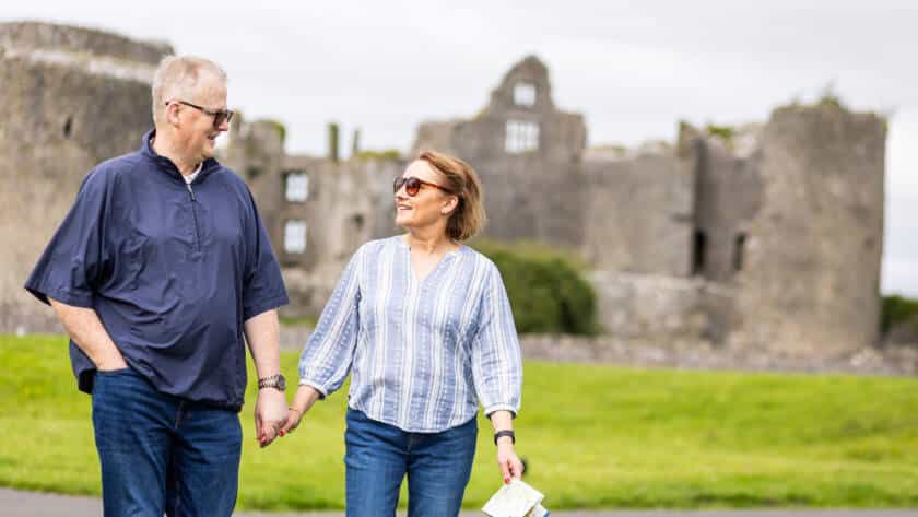 A man and woman, both wearing sunglasses and casual clothes, hold hands and smile at each other whilst walking outdoors. Ancient stone castle ruins are visible in the background on a cloudy day.