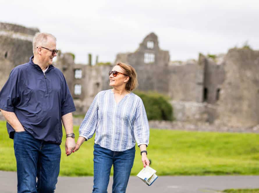 A man and woman, both wearing sunglasses and casual clothes, hold hands and smile at each other whilst walking outdoors. Ancient stone castle ruins are visible in the background on a cloudy day.