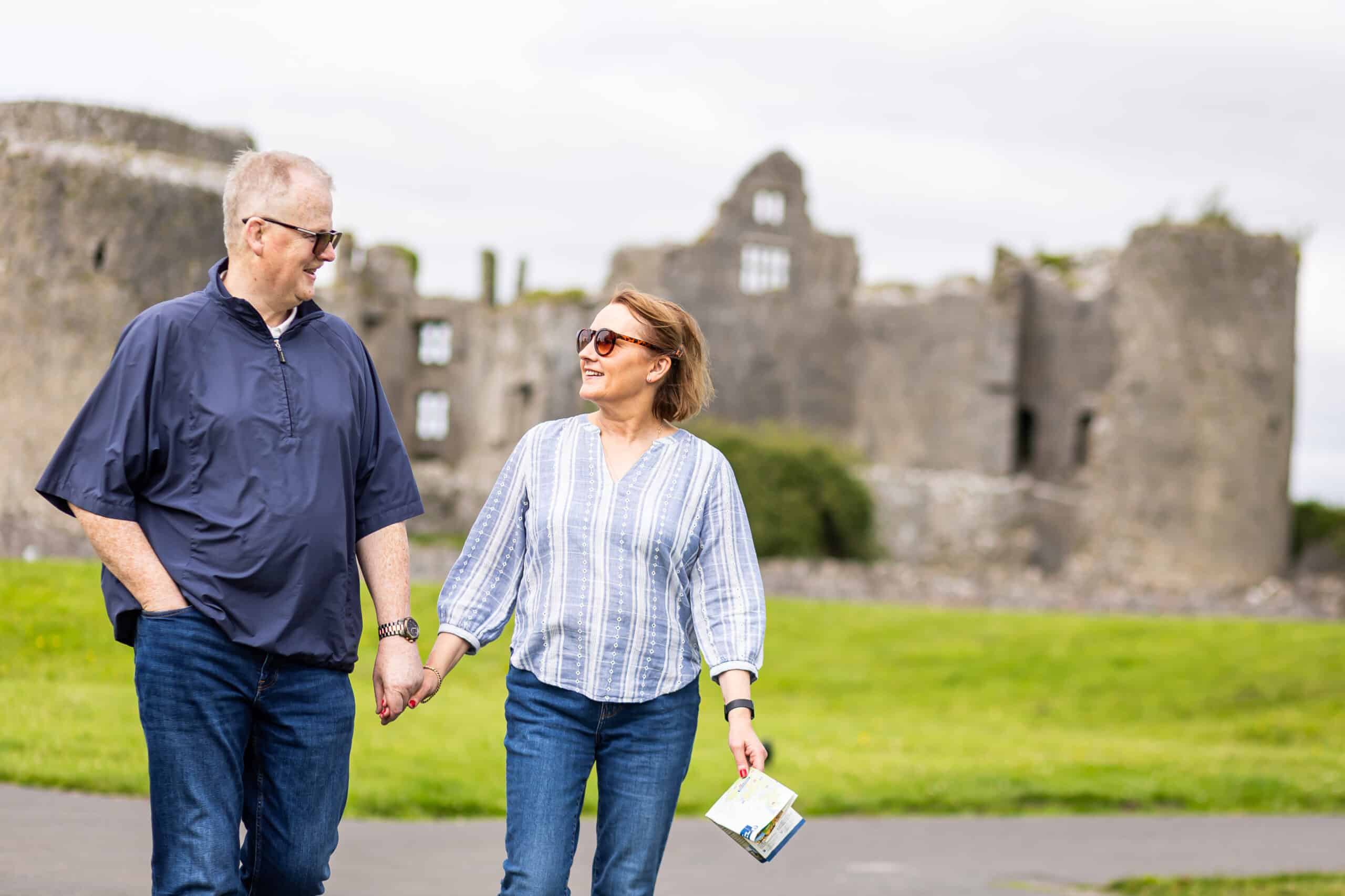 A man and woman, both wearing sunglasses and casual clothes, hold hands and smile at each other whilst walking outdoors. Ancient stone castle ruins are visible in the background on a cloudy day.
