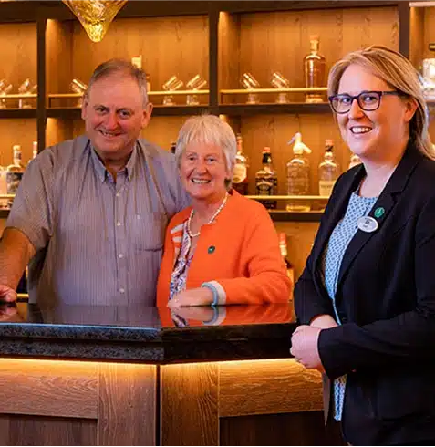 Three people stand smiling at a bar with wooden shelves displaying bottles behind them. Two people stand together behind the counter, while a woman in glasses and a suit stands in front, facing the camera.