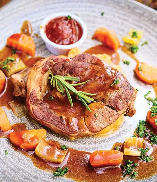 A plate with a cooked meat steak topped with rosemary, surrounded by cooked carrots, onions, and potatoes in brown gravy. A small cup of red sauce is in the background, garnished with chopped herbs.