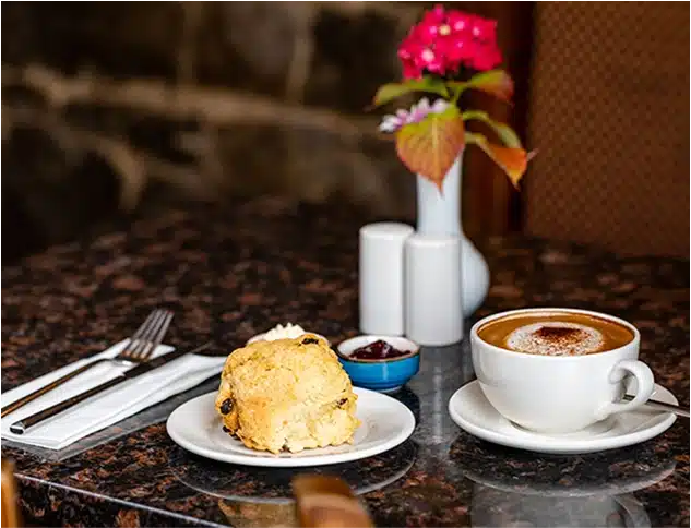 A plate with a scone, jam, and cream sits next to a cup of coffee on a marble table. Nearby are a fork and knife on a serviette, salt and pepper pots, and a vase with a pink flower.