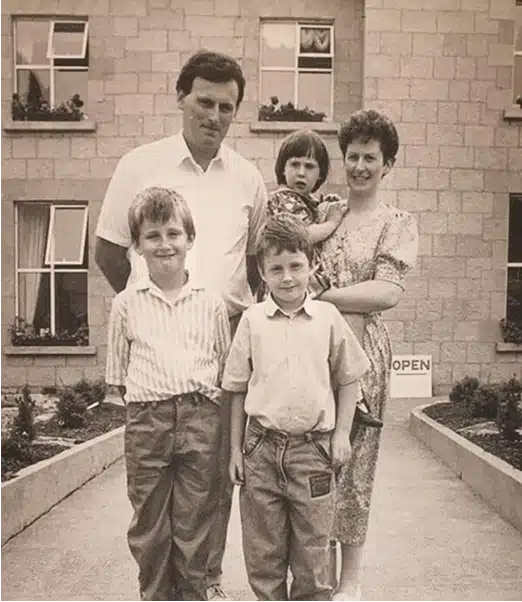 A black-and-white photo of a family of five standing on a path outside a stone building. Two adults stand behind three children, one of whom is being held by the woman. All face the camera and appear to be posing together.