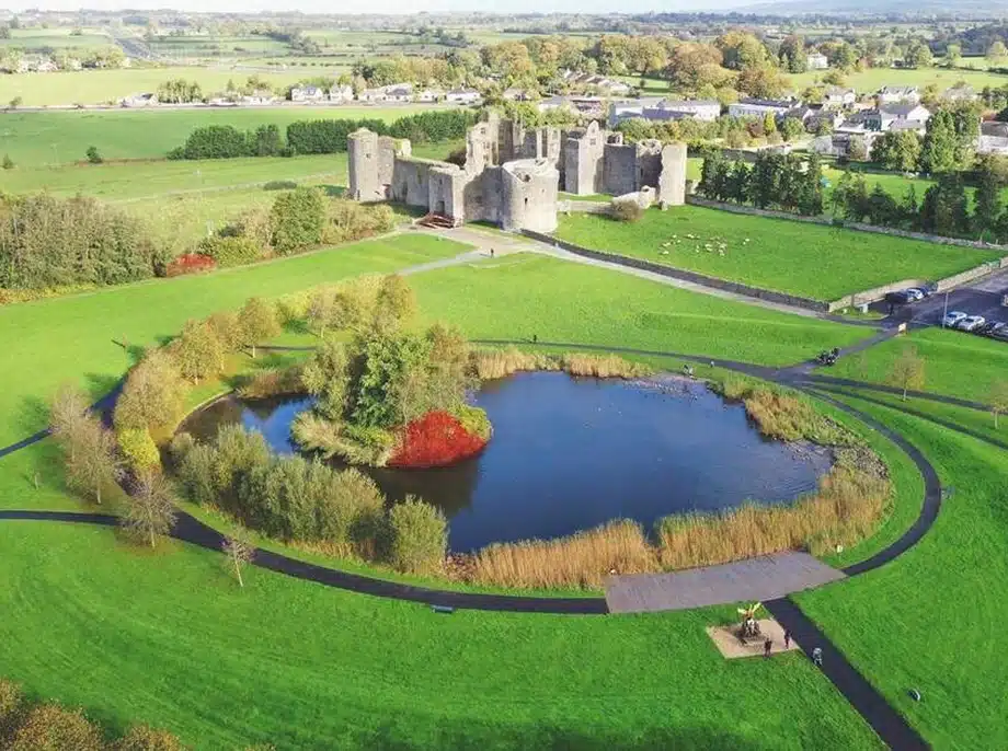 Aerial view of a circular pond surrounded by walking paths and green grass, with trees displaying autumn colours. In the background, there is a large stone castle and a village with fields beyond.