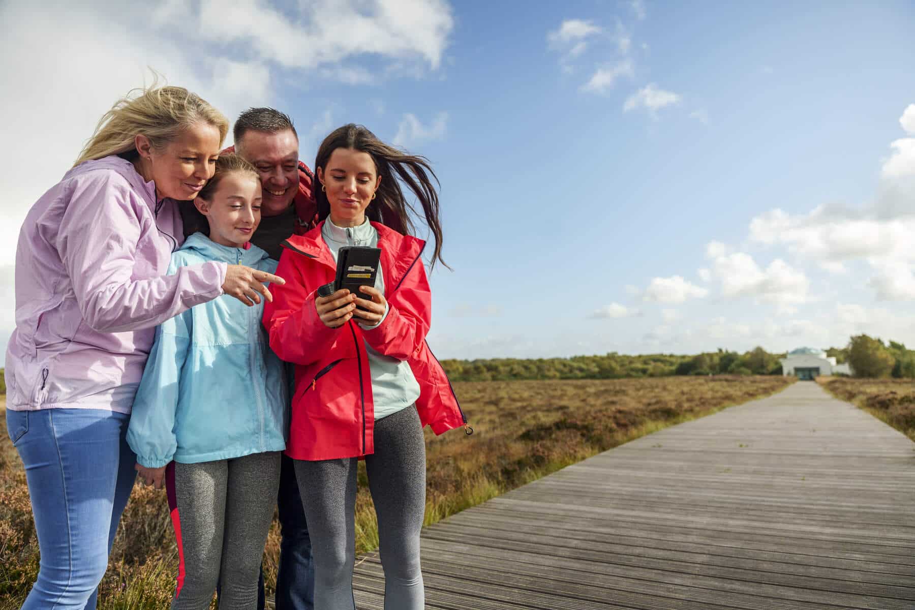 Four people, two adults and two children, stand on a wooden boardwalk outdoors, smiling and looking at a handheld sat nav device together. The weather is sunny with some clouds, and there is greenery around them.