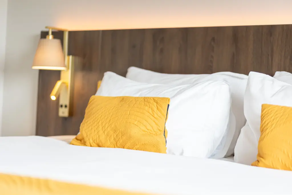 A neatly made bed with white sheets, two white pillows, and two yellow accent cushions, set against a wooden headboard with a modern wall-mounted lamp on the left.