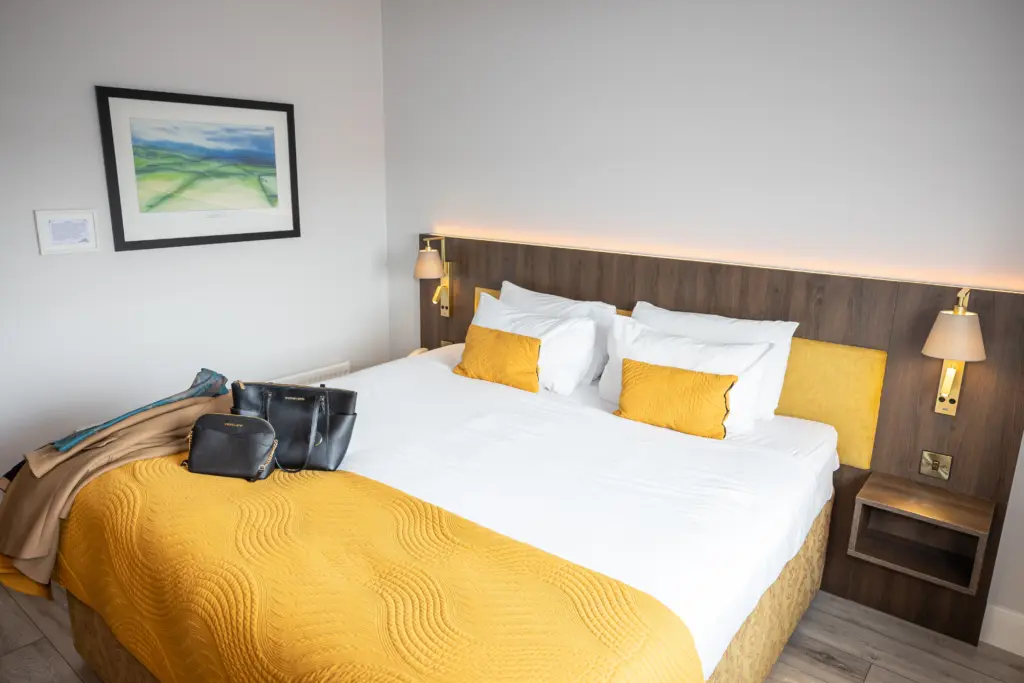 A neatly made bed with a yellow quilt and pillows, a wooden headboard with mounted lamps, and a coat and black bag placed on the bed in a modern, bright hotel room.