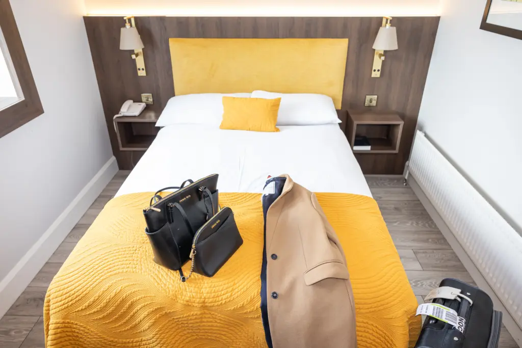 A neatly made bed with a yellow headboard, throw, and pillow. A tan coat, black handbag, and suitcase are placed on the bed in a modern, bright hotel room with wooden flooring.