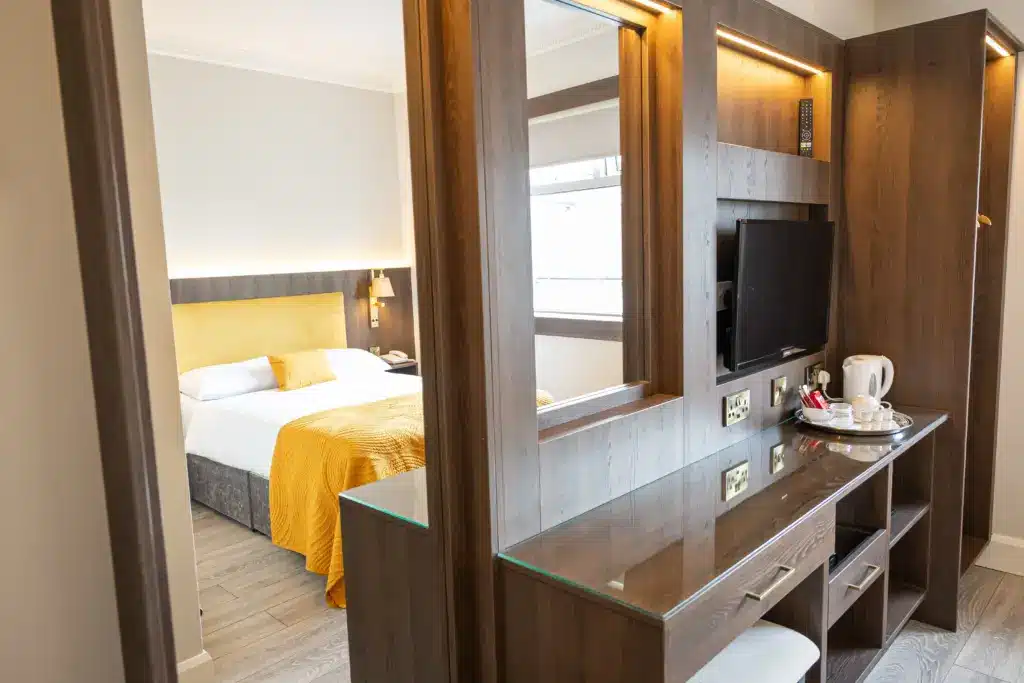 Modern hotel room featuring a wooden desk and mirror, a TV, a tea and coffee set, and a bed with white sheets and a yellow blanket and headboard in the background.