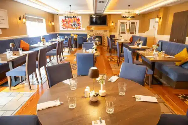 A cosy, well-lit restaurant dining area with blue chairs, wooden tables set with glasses and serviettes, decorative cushions on booth seating, and a fireplace at the back wall beneath a large TV.