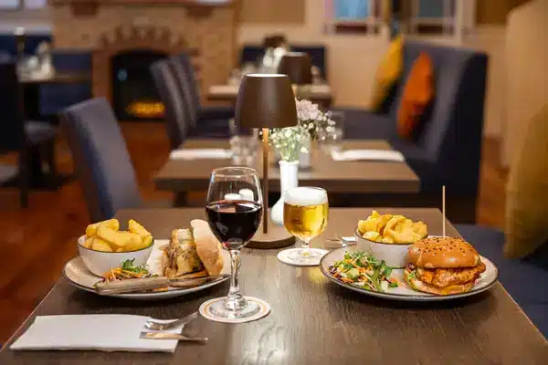 A restaurant table set for two with plates of burgers, chips, salads, a glass of red wine, and a glass of beer. The background shows cosy seating, a lit fireplace, and soft lighting.