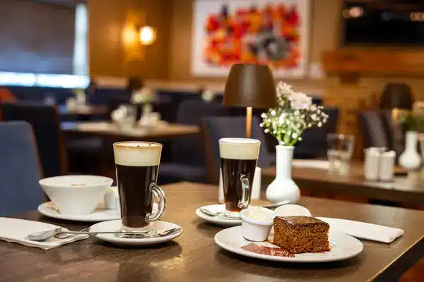 Two glasses of coffee topped with cream, a bowl with a spoon, a plate with cake and cream, and a vase with flowers on a restaurant table; blurred background with more tables and chairs.