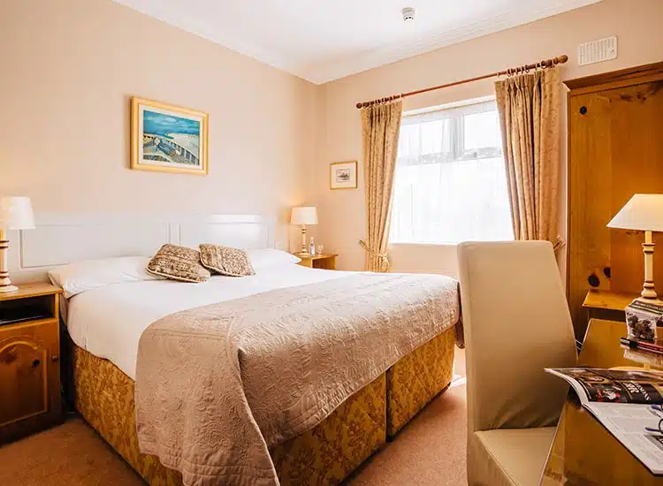 A cosy hotel room with a large double bed, beige bedspread, two bedside tables with lamps, a window with curtains, a wooden wardrobe, a desk and chair, and a painting of a beach scene on the wall.