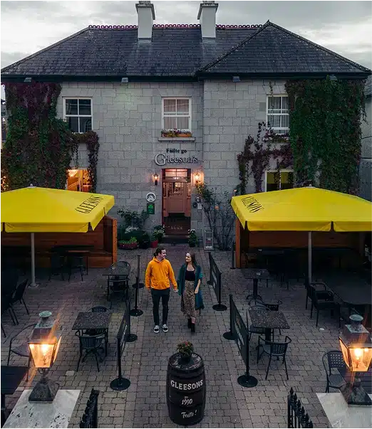 A man and woman walk hand in hand through an outdoor patio with empty tables and yellow umbrellas in front of a stone building labelled “Gleesons.” The building is covered in green and red ivy.