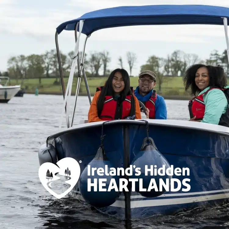 Three people wearing life jackets smile on a small boat in a river, with trees and another boat in the background. The text "Ireland’s Hidden Heartlands" appears with a heart-shaped logo.