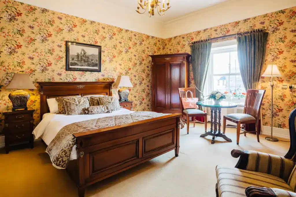A cosy bedroom with floral wallpaper, a wooden bed with patterned bedding, bedside tables with lamps, a wardrobe, two chairs and a small table by a window with curtains, and a framed picture above the bed.
