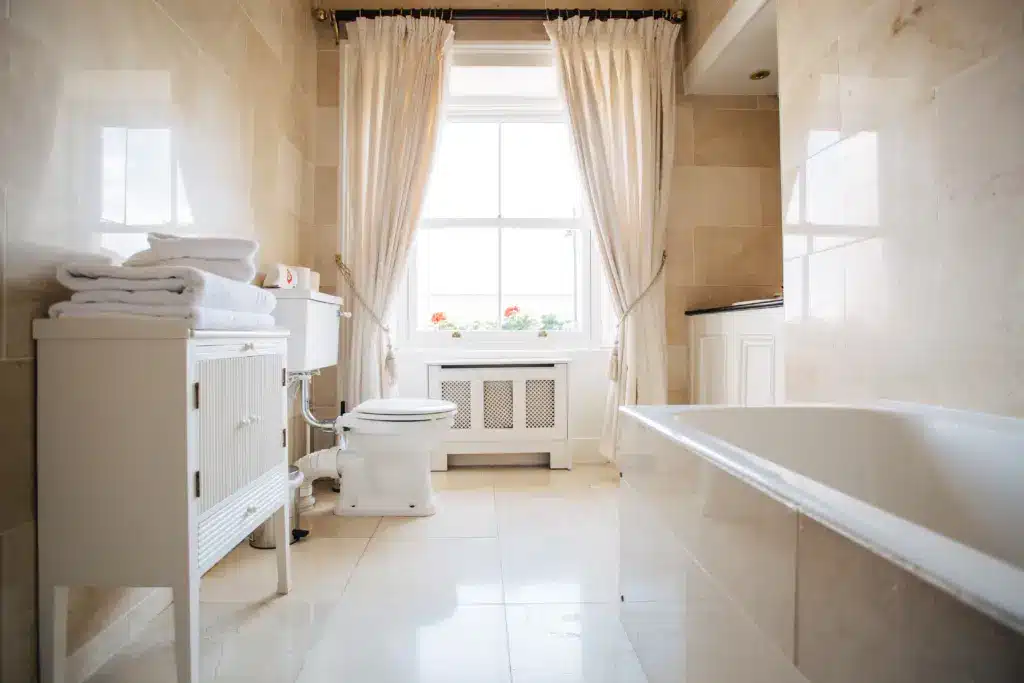 A bright, modern bathroom with beige marble tiles, a white bath, a toilet, a cupboard with folded towels, and large windows with cream curtains letting in natural light.