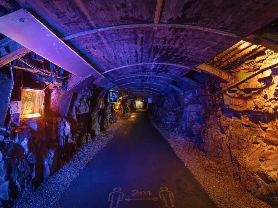 A tunnel with rocky walls and a curved ceiling, illuminated by purple and orange lights, has a sign on the ground indicating a 2-metre distance between people. Signs and gravel line the sides.