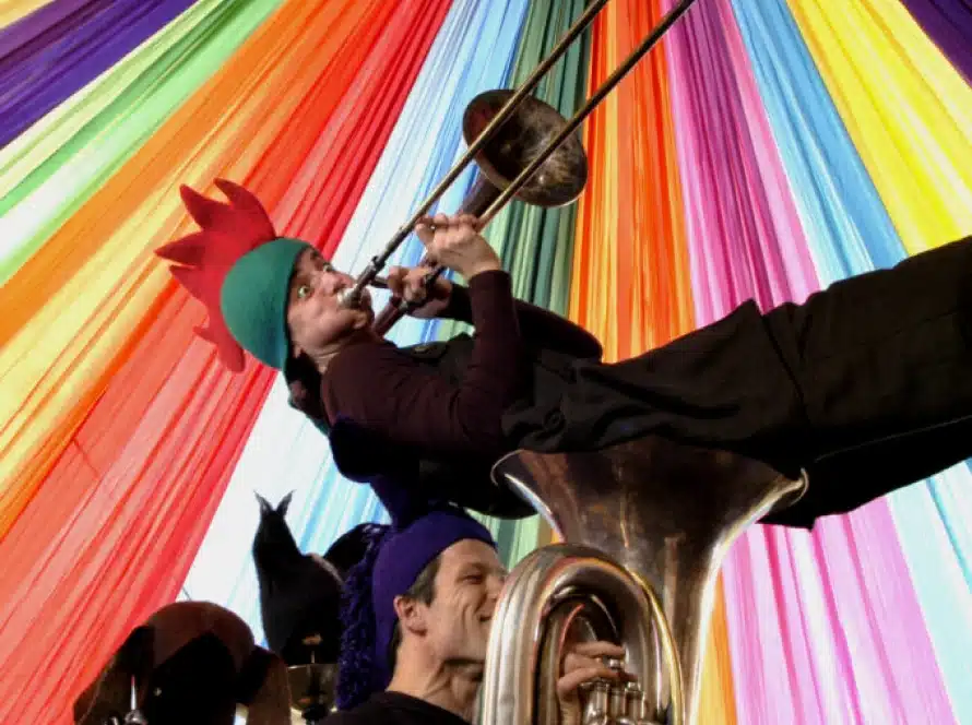 A person in a red hat plays a trombone while leaning back over a tuba player; both are under a colourful, striped marquee ceiling.