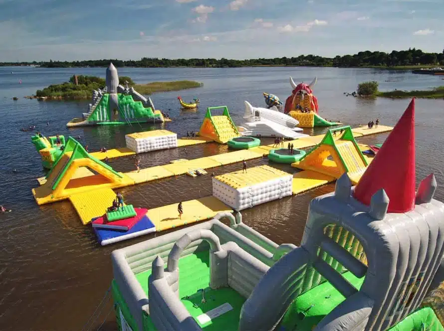 An inflatable water park floating on a lake with slides, climbing obstacles, and platforms in bright colours, surrounded by water and greenery under a partly cloudy sky.