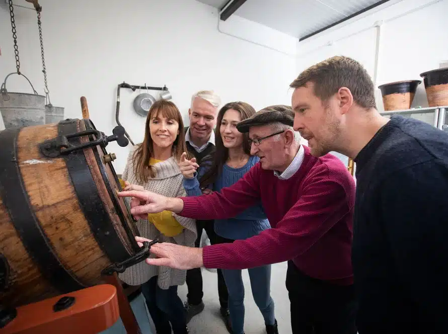 A group of five adults stands around a large wooden barrel, looking inside with curiosity and interest. They are indoors in a well-lit room, appearing engaged and intrigued by what they see.