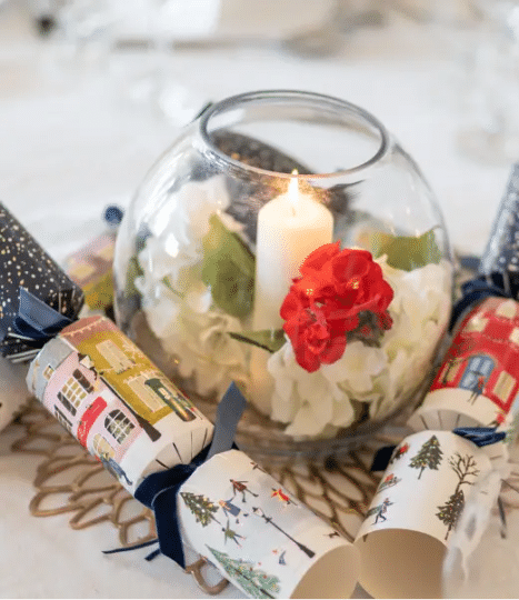 A round glass vase with a lit white candle, white and red artificial flowers inside, surrounded by decorative Christmas crackers with festive designs on a woven mat.