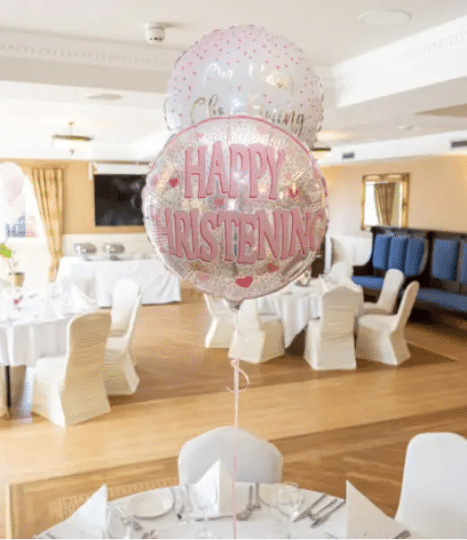 Three pink and white helium balloons with "Happy Christening" text float above a set dining table in a decorated, sunlit function room with white chair covers and wooden floors.