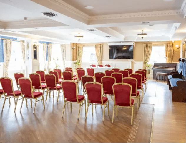 A bright conference room with rows of red cushioned chairs facing a long table, large screen, and a piano by the window; natural light streams in through large windows with beige curtains.