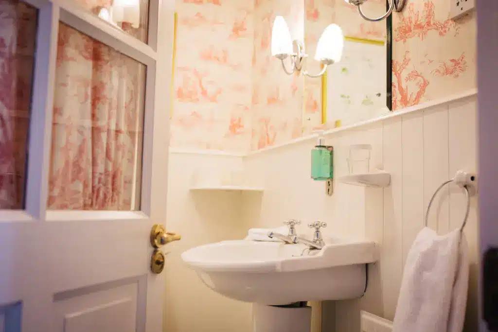 A small bathroom with vintage-style pink and white wallpaper, white panelling, a white basin with chrome taps, a wall-mounted soap dispenser, a white towel, and a light fitting above the mirror.