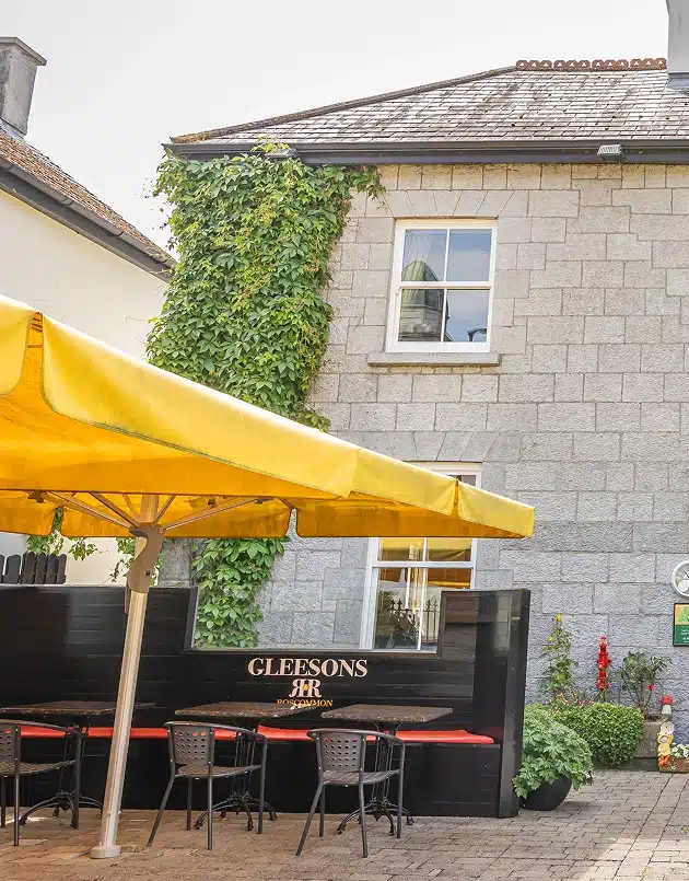 Outdoor seating area with black chairs and red cushions beneath a large yellow umbrella, next to a stone building partially covered in green ivy. The sign “GLEESONS” is displayed on a black panel.