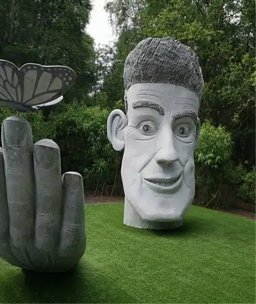 A large, whimsical grey sculpture of a man's smiling face and hand on green grass, with a butterfly perched on the hand, set in a garden with trees and bushes in the background.