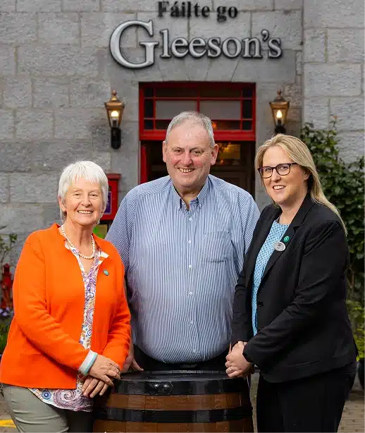 Three adults stand smiling in front of a stone building entrance with a sign that reads "Fáilte go Gleeson's." The two women stand on either side of a man behind a wooden barrel.