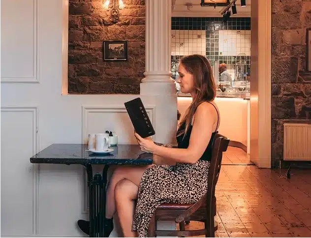 A woman in a patterned skirt sits alone at a café table, reading a menu. A cup and teapot are on the table. The background shows a stone wall, a column, and an open doorway to a kitchen area.
