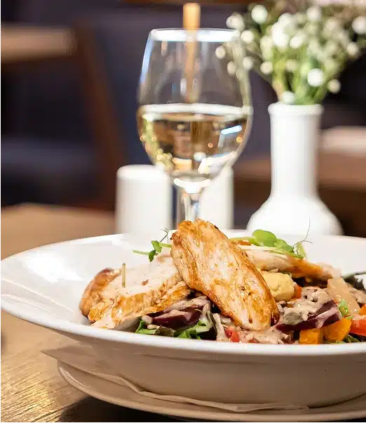 A white plate of grilled chicken salad with mixed leaves, tomatoes, and dressing sits on a wooden table. Behind it, there's a glass of white wine and a white vase with small white flowers.