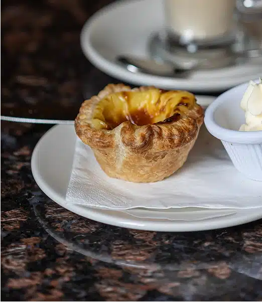 A Portuguese egg tart sits on a white plate with a serviette, next to a small dish of cream, on a dark marble table. A blurred glass and spoon are visible in the background.