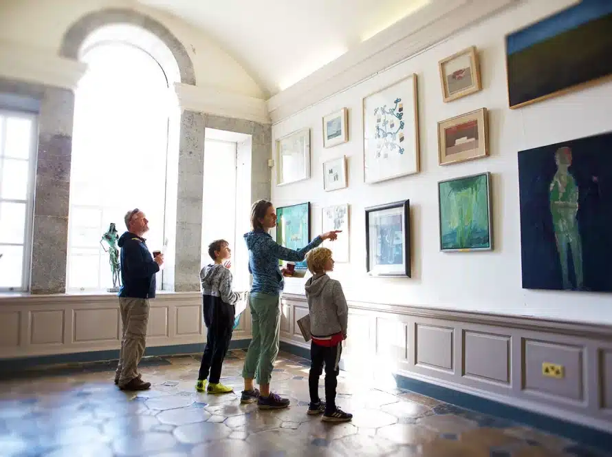 A family of four stands in a bright art gallery, looking at framed artwork on the wall. One adult points towards a painting while two children watch attentively. Sunlight streams through large windows behind them.