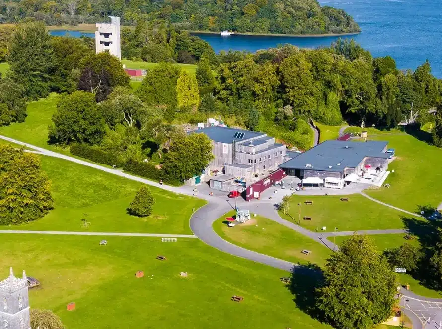 Aerial view of a green park with scattered trees, walking paths, benches, modern buildings, and a lake with forested shoreline in the background.