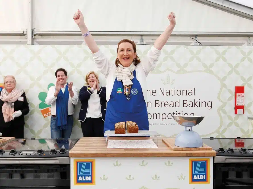 A woman wearing a blue apron stands behind a loaf of bread with her arms raised in victory at a bread baking competition, whilst people behind her clap and smile. Aldi logos are visible on the table.