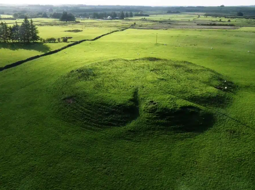 Aerial view of a large, circular grassy mound in a green field, surrounded by stone walls and scattered trees under a clear sky.