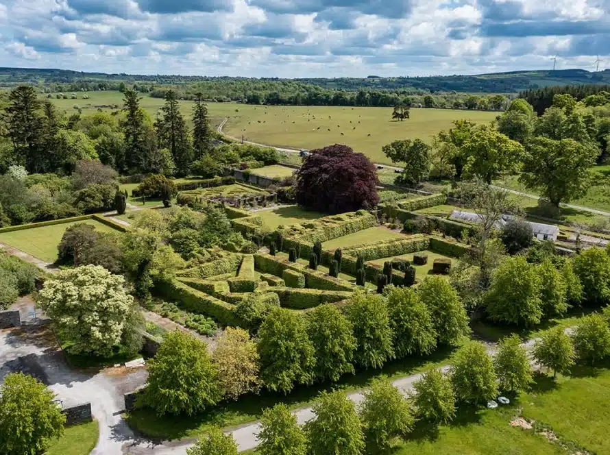 Aerial view of a formal garden with geometrically trimmed hedges, a central large tree, surrounding green lawns, and countryside fields extending into the distance under a partly cloudy sky.