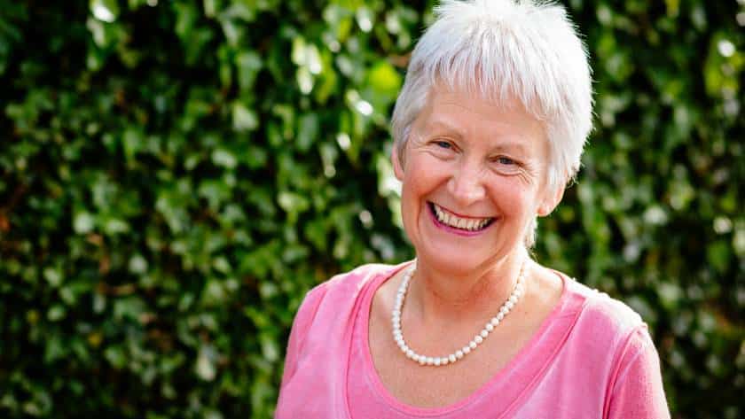An older woman with short white hair, wearing a pink jumper and a pearl necklace, smiles warmly outdoors with a leafy green background.