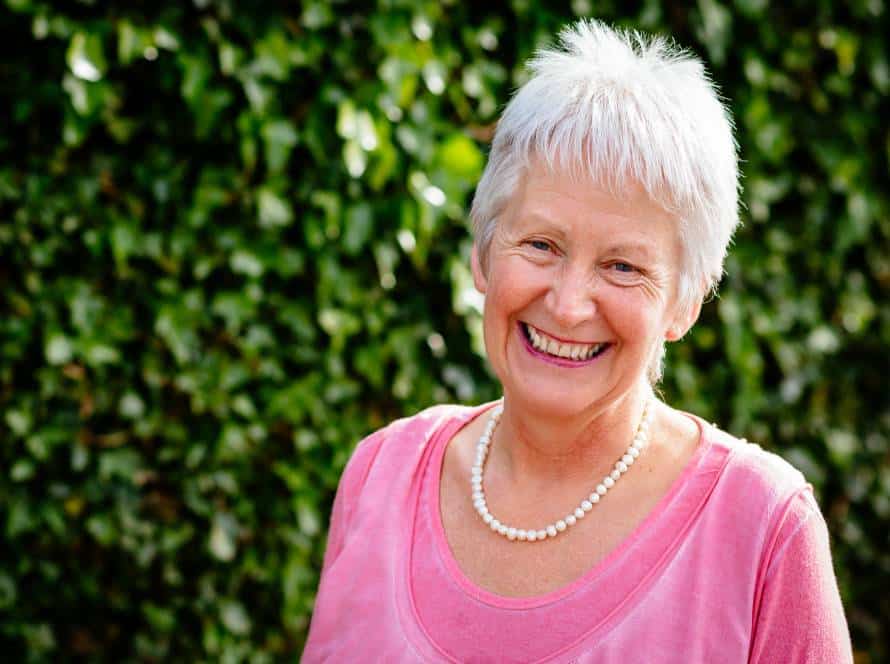 An older woman with short white hair, wearing a pink jumper and a pearl necklace, smiles warmly outdoors with a leafy green background.