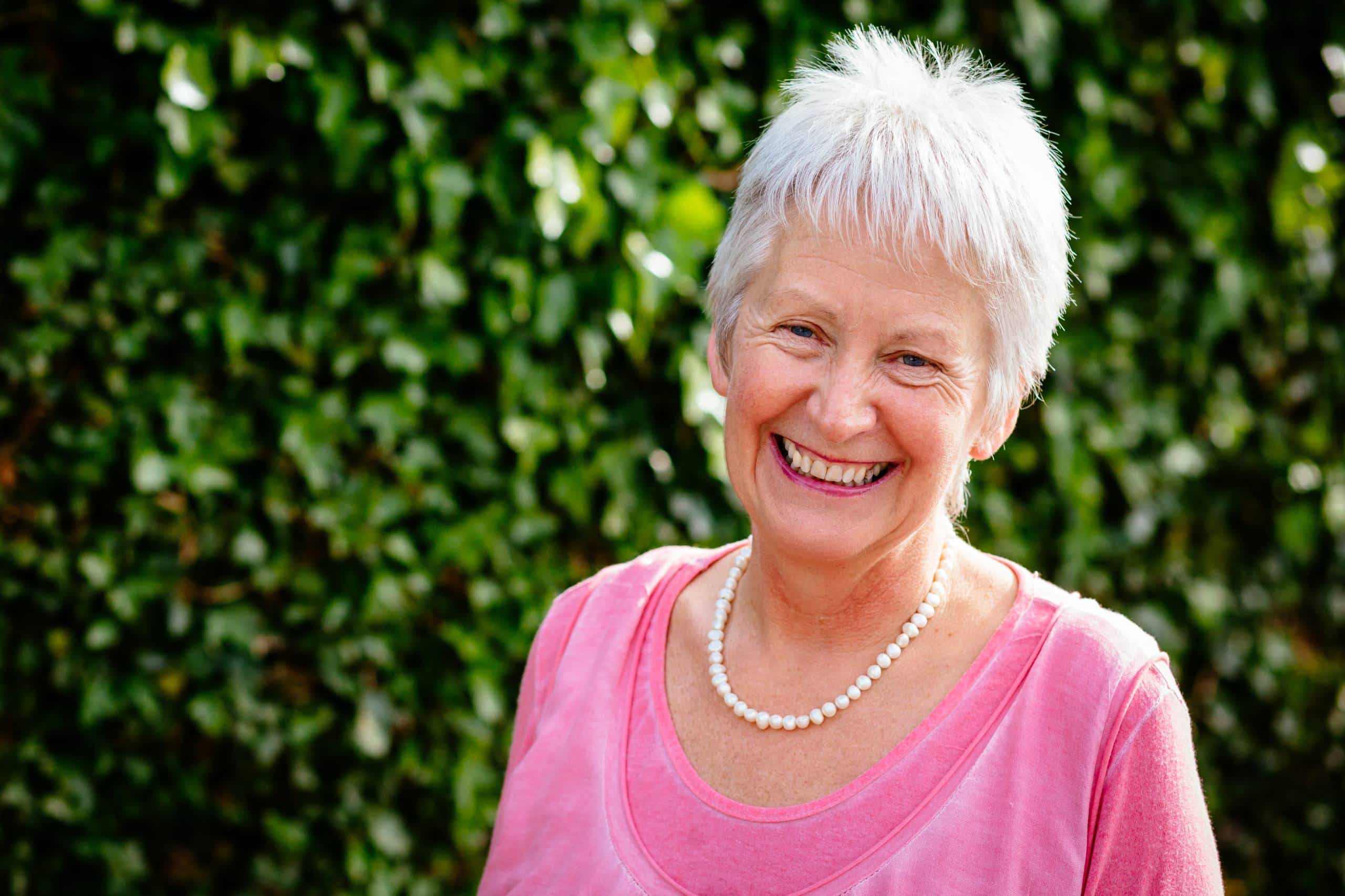 An older woman with short white hair, wearing a pink jumper and a pearl necklace, smiles warmly outdoors with a leafy green background.