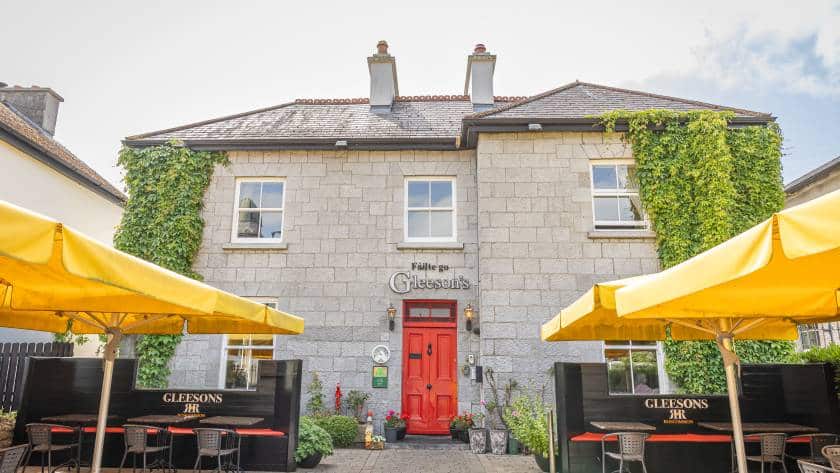 A stone building with ivy on the walls, a bright red door, and yellow patio umbrellas. The building has a sign reading "Gleesons" and outdoor seating areas with plants and flowers.