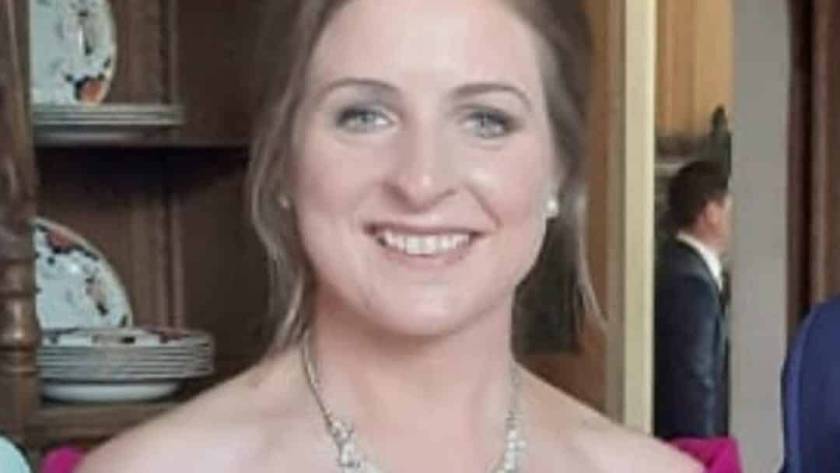 A woman in a bright magenta off-the-shoulder dress stands indoors, smiling at the camera. She wears a silver necklace and her light brown hair is styled up. Shelves with plates and wooden furniture are in the background.