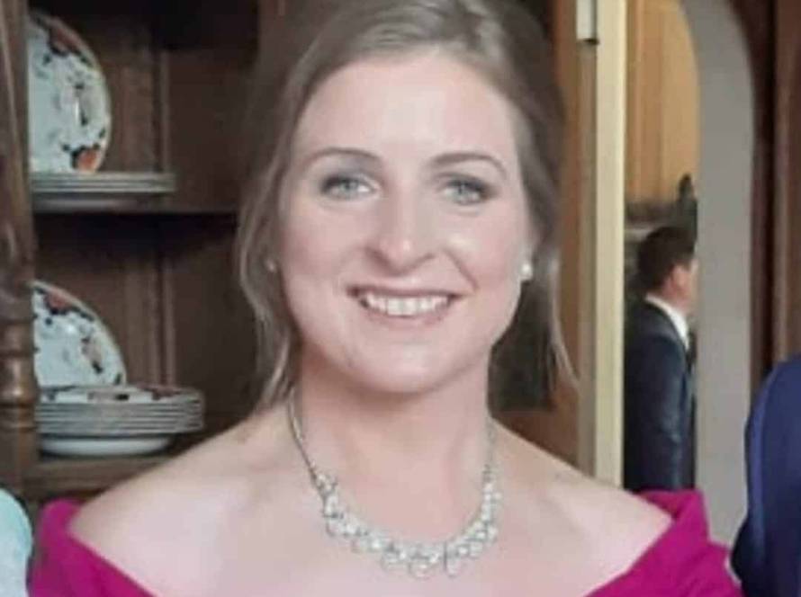 A woman in a bright magenta off-the-shoulder dress stands indoors, smiling at the camera. She wears a silver necklace and her light brown hair is styled up. Shelves with plates and wooden furniture are in the background.
